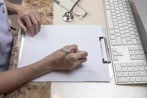 Asian female doctor or nurse is writing something on clipboard with stethoscope and part of computer keyboard on the table at medical office room photo