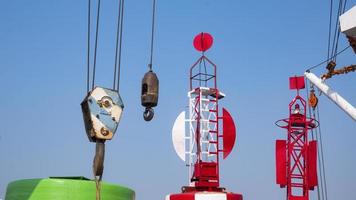 Part of crane hoisting block, hook, steel wire rope with red fairway buoy and white boom against blue sky background photo