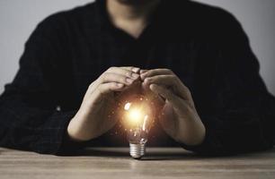 mano de hombre de negocios que protege la bombilla incandescente con un rayo de luz naranja sobre una mesa de madera para inspirar la idea del pensamiento creativo y el concepto de tecnología de innovación futura. foto