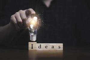 Hand touching to glowing lightbulb on ideas wording which print screen on wooden cube block for creative thinking idea and problem solving concept photo