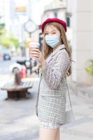 Beautiful Asian woman in long bronze hair who wears grey suit walks in the outdoors of city and wears medical face mask while holding a cup of coffee in hand and looking at the camera in health care. photo