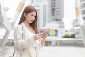 hermosa joven mujer de negocios asiática va a la oficina o al lugar de trabajo que tiene un teléfono inteligente en sus manos en una gran ciudad con edificios comerciales con la ciudad como fondo. foto