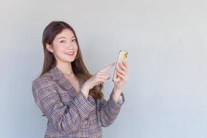 mujer profesional asiática con cabello largo negro usando un traje a cuadros mientras actúa disfrutando con el éxito del trabajo y mirando la cámara usando un teléfono inteligente en un fondo blanco.