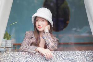 Beautiful Asian woman wears a white hat and plaid coat while she sitting near glassed window and looking out the window in New Year and winter theme. photo