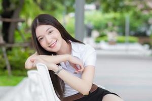 retrato de un estudiante tailandés adulto con uniforme de estudiante universitario. hermosa chica asiática sentada sonriendo felizmente en la universidad con un fondo de árboles de jardín. foto