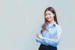 mujer trabajadora asiática profesional que tiene el pelo largo usa camisa azul mientras cruza el brazo y sonríe alegremente sobre fondo blanco.