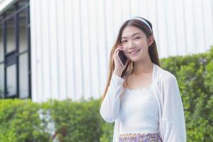 Young Asian business professional woman in white dress is calling phone seriously with someone at outdoor building and bushes as the background. photo