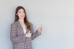 Asian professional woman with black long hair wearing a plaid suit and pretty smiling looking at camera while present product thumbs up means good on white background. photo