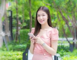 Asian professional  business woman with long hair is smiling in the garden while looking at the camera holding a smartphone in her hand with garden background, work from anywhere concept. photo