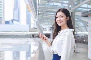 una joven mujer de negocios asiática va a la oficina o al lugar de trabajo donde mira la cámara sostiene un teléfono inteligente en sus manos en una gran ciudad con edificios comerciales con la ciudad como fondo. foto