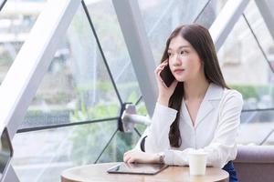 Asian professional business female in a white shirt is while using  your smartphone in your hand to talk she is sitting happy smile working at coffee shop among business background in the city. photo