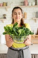 Young brunette smiling woman holding a bowl of fresh spinach in the kitchen photo