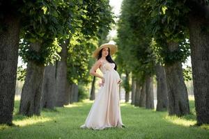 mujer embarazada con un sombrero posando con un vestido sobre un fondo de árboles verdes. foto