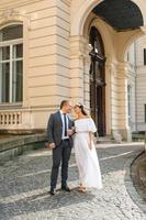 Wedding photo session on the background of the old building. The groom watches his bride posing. Rustic or boho wedding photography.