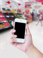 Hand holding mobile phone with Vegetables and fruit on shelf in supermarket blurred background photo