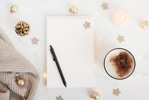 White sheet of notebook with pen on white table with cup of cocoa or coffee, candle and festive garland. Concept planning, wish list for the new year. Mockup for lettering, art drawing. photo