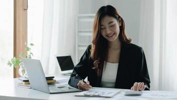 Beautiful Asian woman sitting in office using laptop and calculator. Happy business woman sitting at a desk in an office with a tablet computer. photo