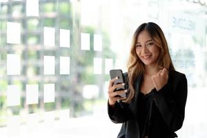 hermosa mujer asiática usando un teléfono inteligente, emocionante y celebrando con buenas noticias de negocios exitosos. concepto de puesta en marcha de pequeñas empresas. foto