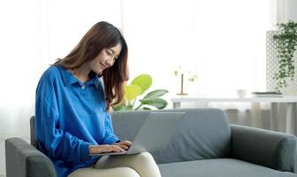 joven mujer asiática independiente de negocios que trabaja en una computadora portátil revisando las redes sociales mientras está acostada en el sofá cuando se relaja en la sala de estar en casa. foto