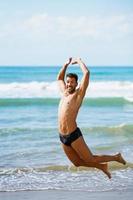 Young man with beautiful body in swimwear jumping on a tropical beach.
