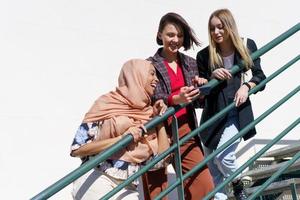 diversas estudiantes alegres sonriendo mientras comparten un teléfono inteligente en la escalera foto