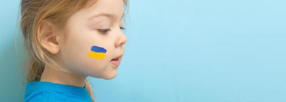 una niña con una bandera ucraniana pintada de amarillo y azul en las mejillas. copie el espacio sobre fondo azul foto