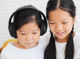 Adorable two little kid sisters enjoy watching online movies or playing games on wireless tablet computer. A cute child with a headset and her sister relaxing at home concentrates on gaming together. photo