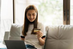 Online shopping concept. Dreamy asian woman with computer laptop and credit card sitting on couch, thinking about internet purchases or food delivery, free space. photo