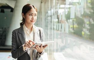 Asian woman with digital tablet standing in modern office background and copy space, Fashion business photo of beautiful girl in casual suit with tablet.