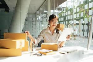 Retrato joven y atractiva propietaria de asia empresa de inicio mira el trabajo de la cámara feliz con la caja en casa prepara la entrega de paquetes en la cadena de suministro de pymes, compras, concepto de comercio omnicanal en línea. foto