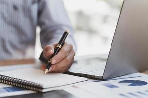 Businessman hand are taking notes on paper with a black pen, and using a laptop computer on a white desk in the office. photo