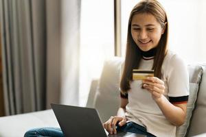 Online shopping concept. Dreamy asian woman with computer laptop and credit card sitting on couch, thinking about internet purchases or food delivery, free space. photo