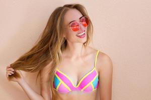 cerca de una chica feliz con gafas de sol rosas aisladas. vacaciones de verano y fin de semana divertido. concepto de verano. mujer joven sonriente en traje de baño de moda. enfoque selectivo. traje de playa de verano. copie el espacio foto