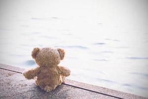 A sad teddy bear sits on a bridge alone looking at the sea in lonely with copy space. Toy, doll, for child.