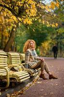 Beautiful red-haired girl with curly hair and blue eyes. The girl is wearing a checkered jacket. The girl is sitting on a bench. photo