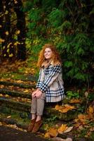 Beautiful red-haired girl with curly hair and blue eyes. The girl is wearing a checkered jacket. The girl is sitting on a bench.