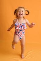 una niña vestida con traje de baño a la edad de un año y medio está saltando o bailando. la niña es muy feliz. foto tomada en el estudio sobre un fondo amarillo.