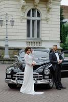 elegante novia hermosa y novio guapo abrazándose en un elegante coche negro a la luz. vista inusual desde atrás. pareja de bodas de lujo en estilo retro. foto