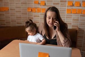 Young beautiful mother is trying to work remotely. Her little daughter is bothering her. photo