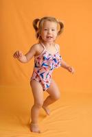 una niña vestida con traje de baño a la edad de un año y medio está saltando o bailando. la niña es muy feliz. foto tomada en el estudio sobre un fondo amarillo.