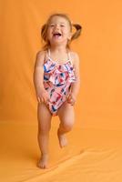 A little girl dressed in a swimsuit at the age of one and a half years is jumping or dancing. The girl is very happy. Picture taken in the studio on a yellow background.