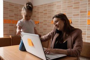 Young beautiful mother is trying to work remotely. Her little daughter is bothering her. photo