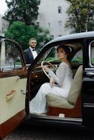 Beautiful happy young bride and groom looking from retro auto photo