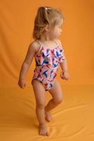 una niña vestida con traje de baño a la edad de un año y medio está saltando o bailando. la niña es muy feliz. foto tomada en el estudio sobre un fondo amarillo.