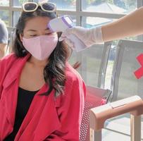 Vertical image of young asian female traveler wearing face mask, sitting follow social distancing regulation and make screening test for COVID-19 by airport staff. New normal travel lifestyle concept. photo