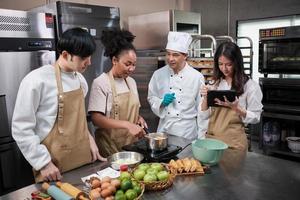 Hobby cuisine course, senior male chef in cook uniform teaches young cooking class students to prepare, mix and stir ingredients for pastry foods, fruit pies in restaurant stainless steel kitchen. photo