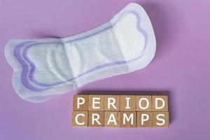 Period cramps text on wooden block. Sanitary pads on purple background. Top view. photo