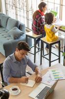 Mixed race family stays at home together, father works at desk with graph and charts and a laptop notebook computer while mother working and teaching little girl in blur background photo