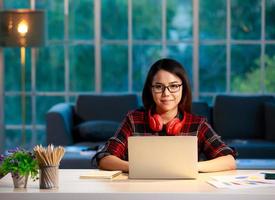 Cute Asian businesswoman working at home living room with twilight sky in outside of house background. She works with laptop notebook computer and looking to camera photo