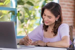 The beautiful middle-aged woman sitting in food shop and working with laptop note computer and writing on memo paper. Freelancer job and work from anywhere concept photo